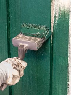 En hånd iført en malehanske holder en pensel som maler en grønn dør med frisk grønn maling.