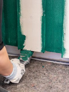 En person maler en hvit kledning grønn med en pensel, bilde viser bare hånden til personen.