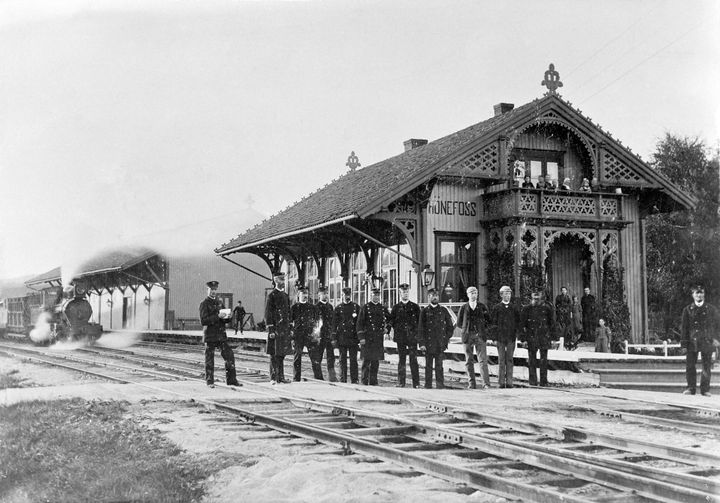 Personalet på Hønefoss stasjon i 1896 med stasjonsbygningen og tog 184 "Slaven" i bakgrunnen. Foto: H. Carlsen/ Norsk Jernbanemuseum