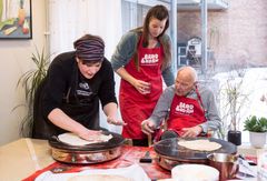 Arnfinn Nordbø, sammen med barnebarnet Ragnhild Blomstrøm Nordbø, følger nøye med og kommenterer mens Ingrid Lamark fra Norges bygdekvinnelag sørger for nybakte lefser til beboerne på Midtåsenhjemmet. Foto: brodogkorn.no