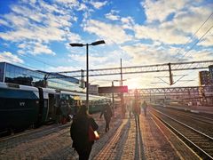 Lav sol over perrongene på Oslo S. Folk går bortover plattformen, mot sola, mens et tog står ved ett av sporene.