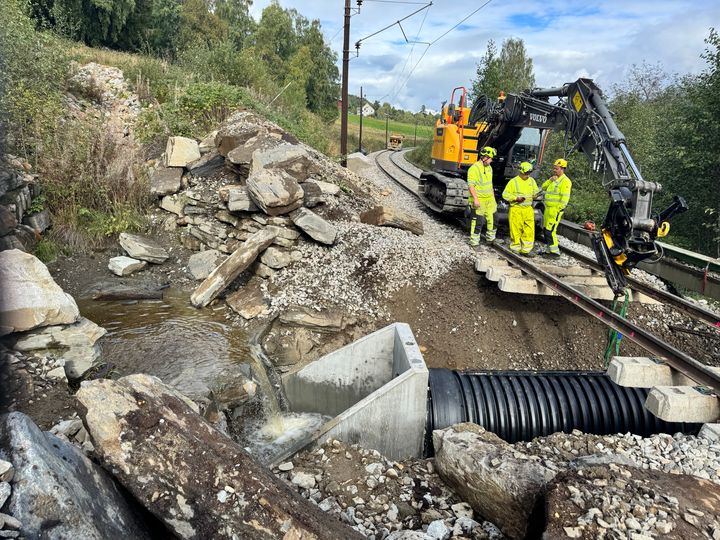 3 menn i gule arbeidsklær og arbeidsmaskin legger stort rør under jernbaneskinner.