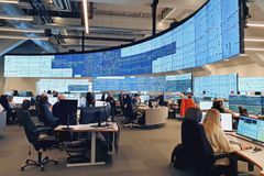 Mange mennesker jobber foran PC. En stor skjerm med masse data henger også på veggen foran dem.