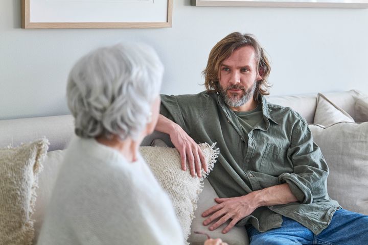 Mann i 40-årene snakker med kvinne i 70-årene i en sofa. Vi ser ansiktet til mannen, men kvinnen sitter med ryggen til.