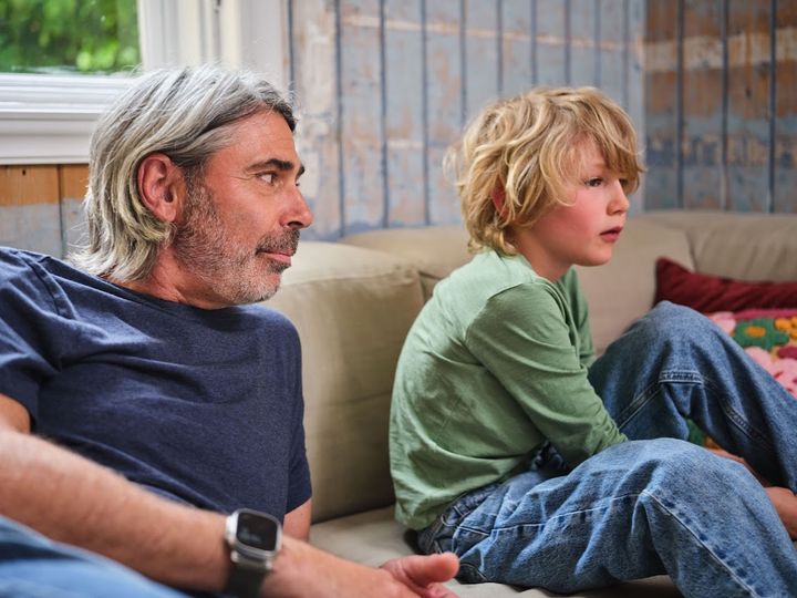 Far og sønn sitter sammen i en sofa. Far ser på sønnen sin, som ser ut i lufta.