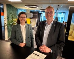 To personer smiler og står ved et bord i et kontormiljø.