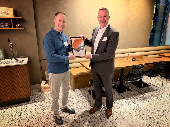 To menn står inne i et moderne rom, og holder et diplom mellom seg.