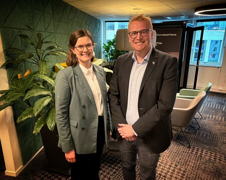 To personer i et kontormiljø, står ved siden av hverandre og smiler.