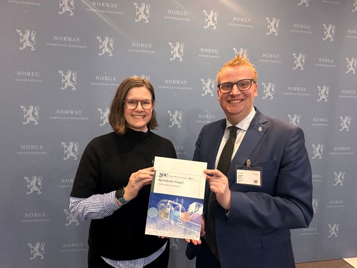 Kvinne og mann holder en fersk NOU mellom seg mens de smiler mot kamera.