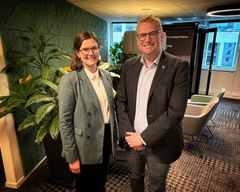 Alt-tekst: En kvinne i grå dressjakke og en mann i mørk dressjakke står og smiler ved siden av hverandre i et kontorlokale med planter og lenestoler i bakgrunnen.