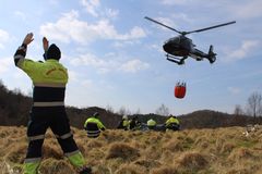 Brannmannskap signalerer til helikopter som bærer en vannbøtte under øvelse i gresslandskap.