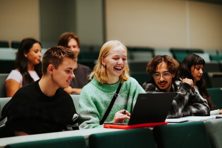 Studenter sitter sammen i et klasserom, smilende over en laptop.