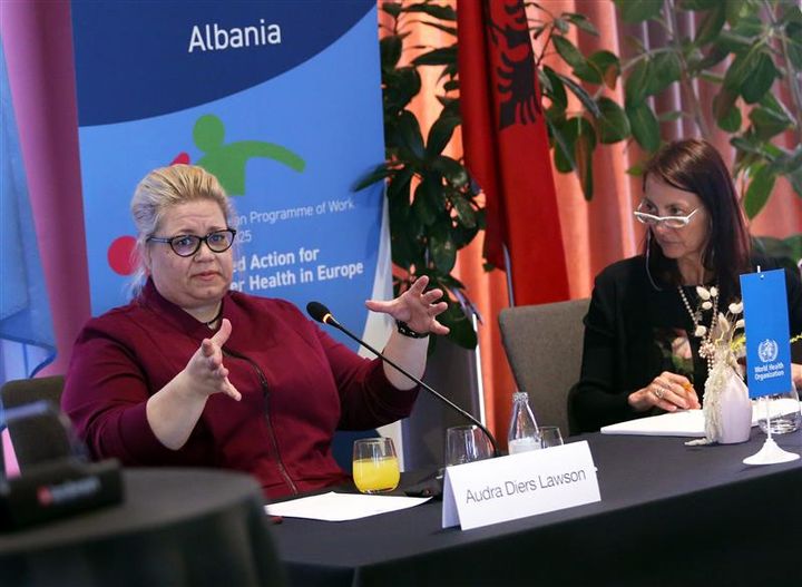 To personer sitter ved et bord med mikrofoner, foran et banner med teksten "Albania".