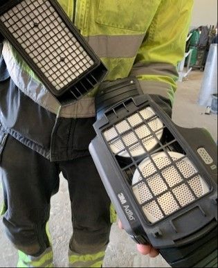 Bildet viser en person som holder frem et motorassistert luftforsynt åndedrettsvern. Verneutstyret er åpent slik at man ser filtrene.