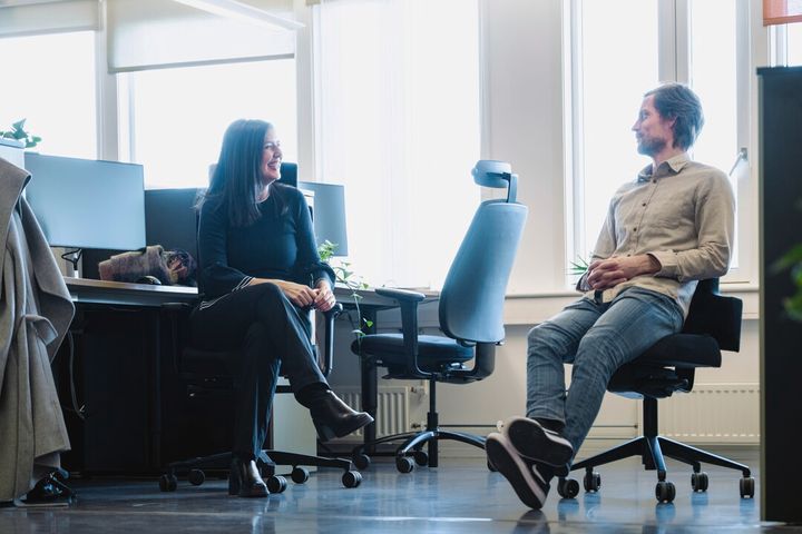 Bildet viser en mann og ei kvinne som sitter i et åpent kontorlandskap og snakker sammen.