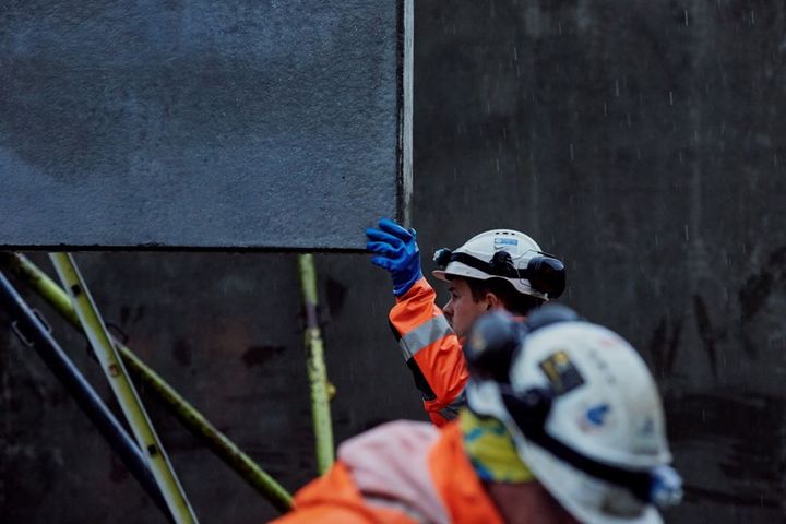 Bildet viser to arbeidere på en byggeplass. Den ene personen tar imot ei bygningsplate som senkes ned med ei kran.