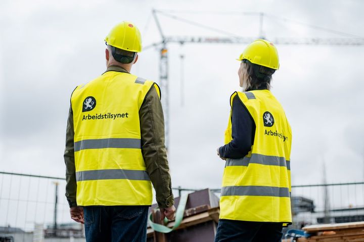 Bildet viser to inspektører fra Arbeidstilsynet med gule vester. Inspektørene står foran en byggeplass med ryggen mot kameraet.