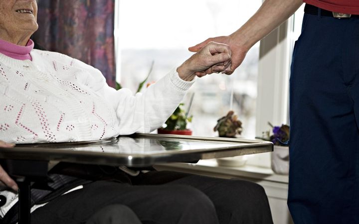 Utsnitt av et fotografi som viser munnen og halsen til en gammel dame som sitter. Og en person som står som holder i hånden hennes.