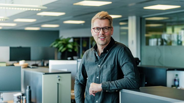 Bildet viser en mann som står i et kontorlandskap. Mannen smiler til kameraet.