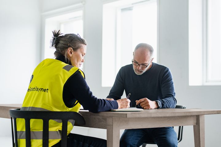 Bildet viser to personer som sitter ved et bord. Den ene har på seg gul Arbeidstilsynet-vest.