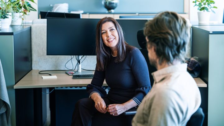 Bildet viser to kolleger, ei dame og en mann, som snakker med hverandre i et åpent kontorlandskap.