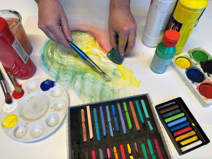 Bilde viser to hender som maler, med pensel i den ene hånden og klut i den andre. På bordet ligger maling, vannfarger og fargestifter.