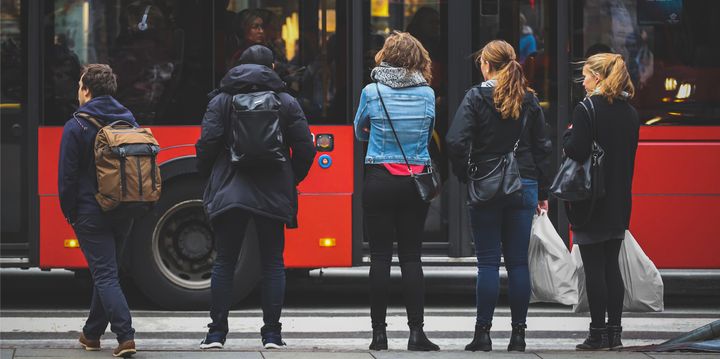 Fem personer står på et fortau. Forbi dem kjører en rød buss.