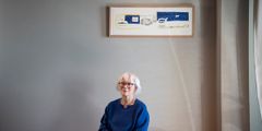 Foto av Britta Marakatt-Labba som sitter på en benk, og på veggen bak henne henger et av hennes kunstverk.