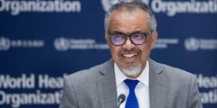 Foto av en smilende Tedros Adhanom Ghebreyesus som står på en talerstol foran en bakvegg med WHO-logoer.