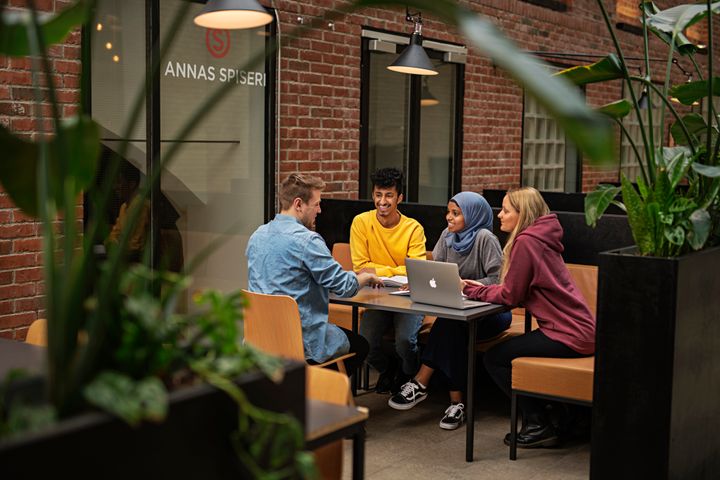 En gruppe på fire personer sitter rundt et bord og snakker, med en bærbar datamaskin foran seg.