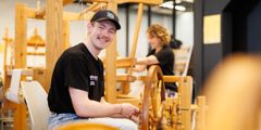 Dennis Dahle Karlsen sitter ved rokken og spinner. I bakgrunnen ser vi Gina Therese Stene veve.