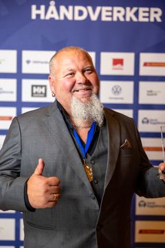 Gothard Høyen Østerhaug smiler og viser tommelen opp foran sponsorveggen på finalen av Norges Hyggeligste Håndverker 2025.