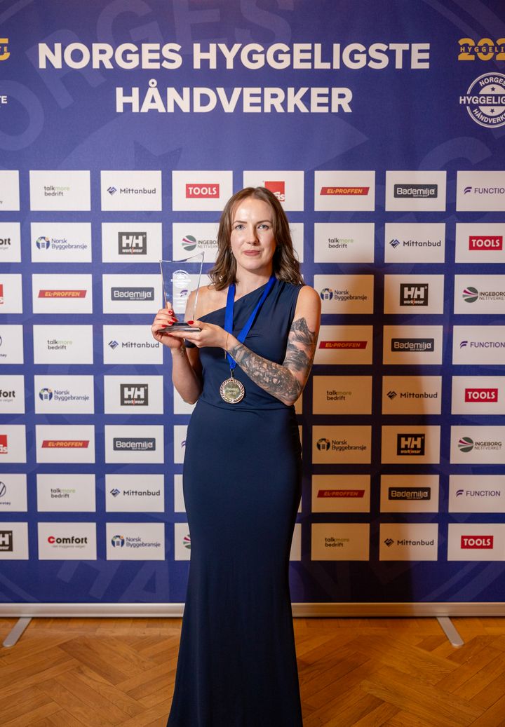 Ewelina Sekiw står foran sponsorveggen med pokalen i hånda under kåringen av Norges Hyggeligste Håndverker 2025