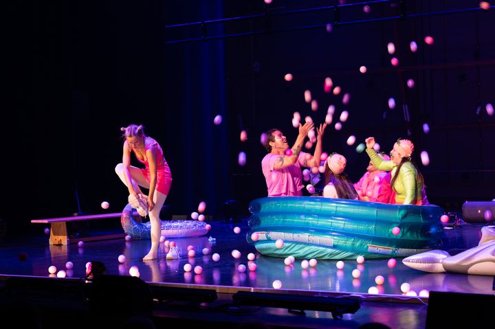 Fire personer sitter i et oppblåsbart badebasseng på en scene og kaster baller i opp i lufta, mens en person står ved siden av og tar på seg skoene.