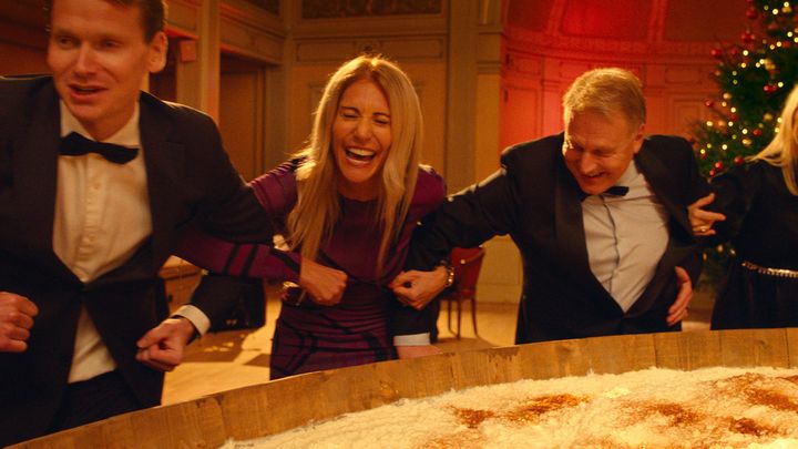 To menn og en kvinne går arm i arm rundt en stor skål med julegrøt, mens de ler og har det gøy.