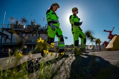 To personer i arbeidsklær går bortpoer en vei med sol og et anleggsområde i bakgrunnen.