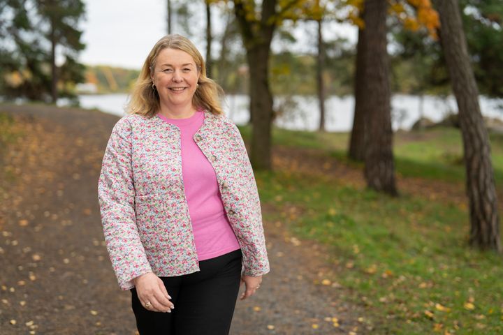 Smilende kvinne gående på en sti. I bakgrunnen ser man gress, trær og oransje blader.