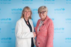 Påtroppende styreleder for UNICEF Norge, Bente Lier i hvit jakke til venstre, og avtroppende styreleder Hilde Britt Mellbye smiler og håndhilser foran en blå UNICEF-bakgrunn.