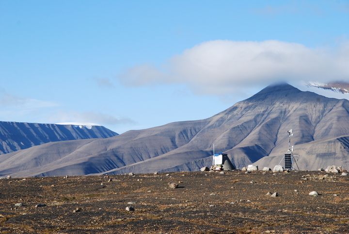 Bildet viser en kjegleformet betongkloss som er målestasjonen for permafost, til høyre vises en værstasjon. Disse er plassert innerst i en dal. Bak skimtes fjell.