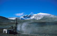 Fjell og tundra på Dovrefjell, snødekket Snøhetta ruver i horisonten, boremaskin og røyk fra boremaskin i forgrunnen.