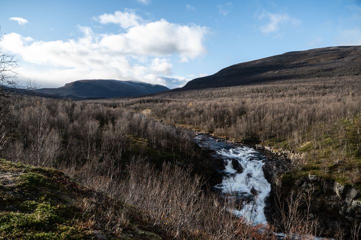 blå himmel med noen skyer, en elv som renner i en dal, brune trær uten løv, brune og butte fjelltopper