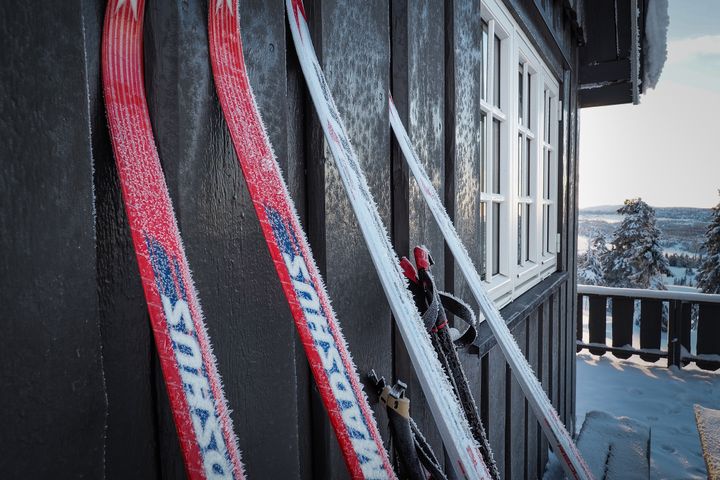 To par ski står lent mot en hyttevegg ved vinduet, med snødekt landskap i bakgrunnen.