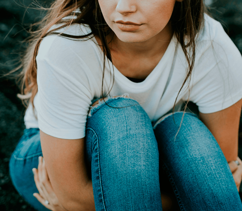 Illustrasjonsfoto: Ung jente sitter på bakken og holder seg rundt knærne. Deler av ansiktet synes. Langt hår, hvit t-skjorte og olabukser.