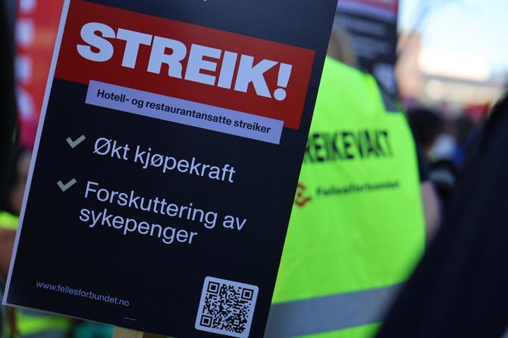 Skilt med teksten "Streik! Hotell- og restaurantansatte streiker. Økt kjøpekraft. Forskuttering av sykepenger." Personer med gule vester i bakgrunnen.