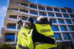 To personer i gule vester med "streikevakt"-tekst står med ryggen til foran en kontorbygning, hever knyttnevene i været.