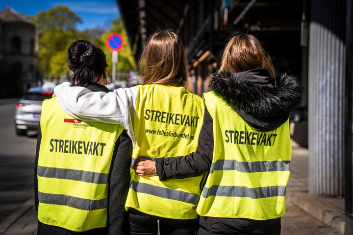 Tre personer med gule vester merket "STREIKEVAKT" står sammen på en gate.