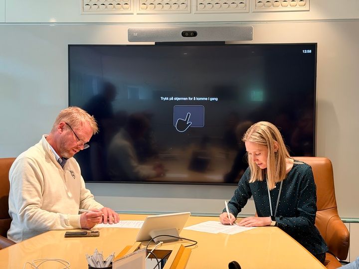 To personer signerer dokumenter ved et bord, med en stor skjerm bak seg.