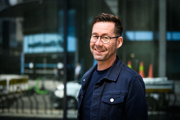 En smilende mann med briller og kort hår, iført en mørk jakke, står utendørs.