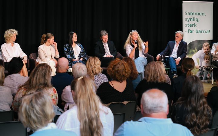 En gruppe mennesker sitter på scenen og deltar i en paneldiskusjon foran et publikum. En plakat i bakgrunnen har teksten: "Sammen, synlig, sterk - for barna!".