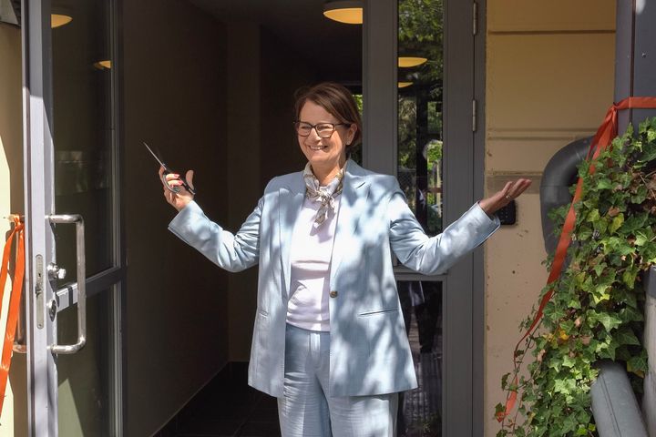 Kvinne i blå dress smiler og holder en saks ved inngangen til en bygning med klippet snor.